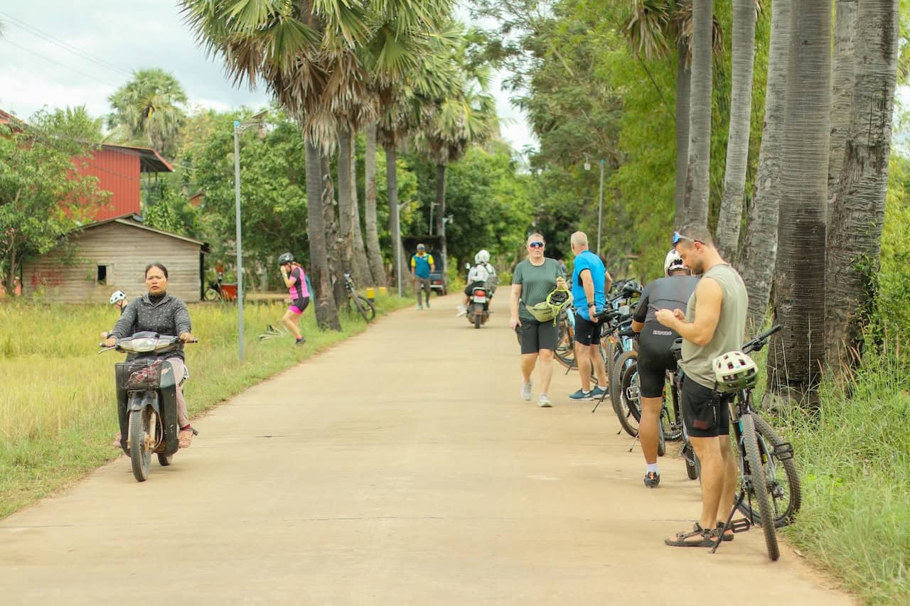 Cyclists riding through rural Cambodian villages between Siem Reap and Phnom Penh, passing rice fields, traditional Khmer houses, local markets, and countryside backroads.