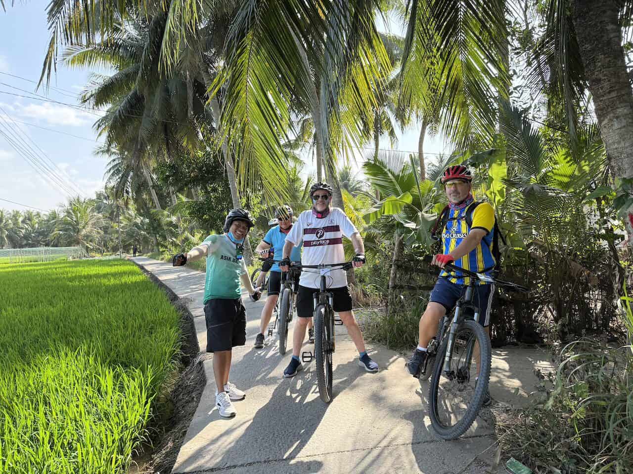 Cyclists riding from Ho Chi Minh City into the Mekong Delta, passing riverside villages, coconut groves, rice fields, and peaceful countryside roads on a Vietnam rural bike journey.