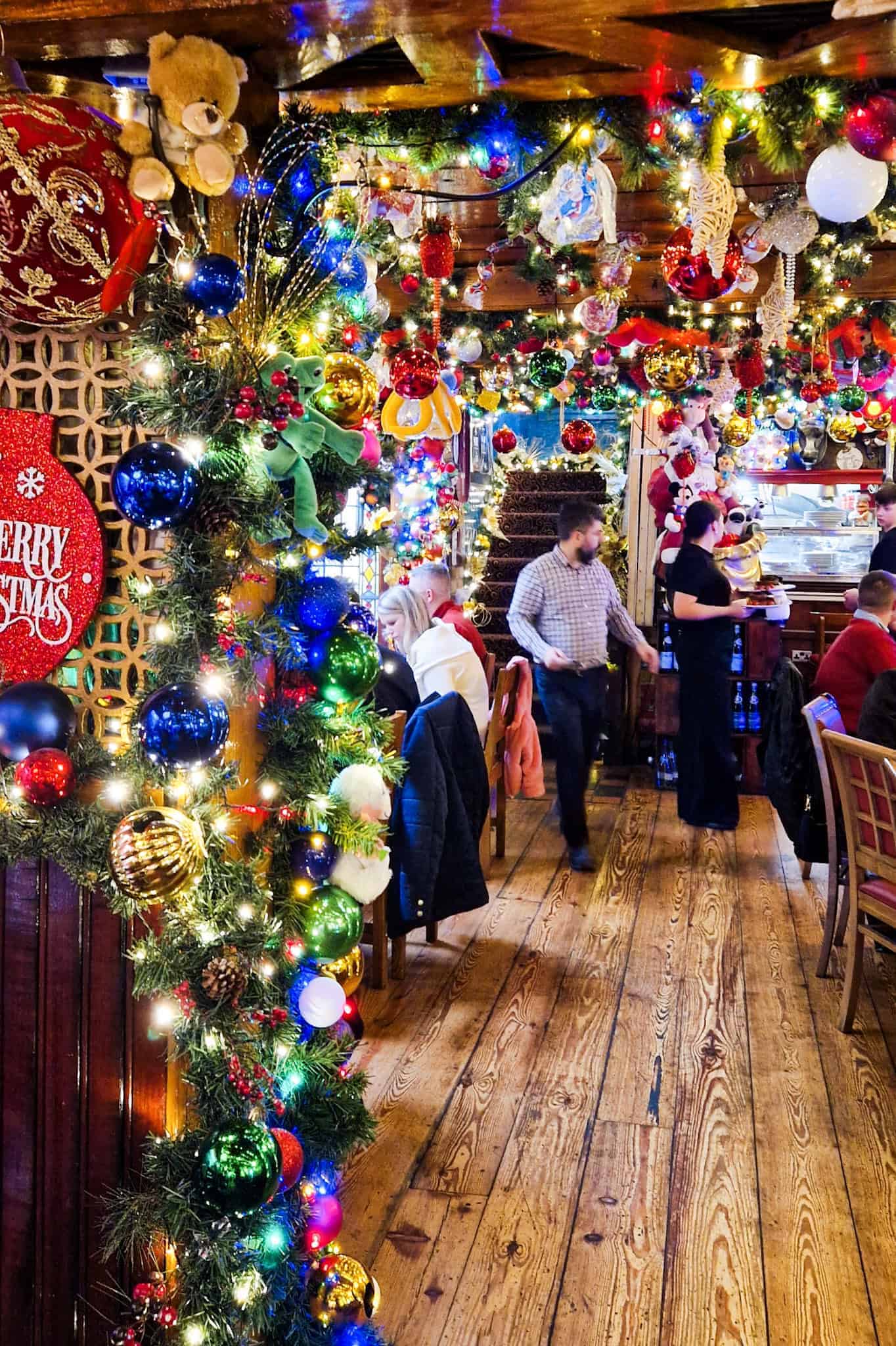 A festive interior of a restaurant decorated with bright Christmas lights, colorful ornaments, and garlands, featuring teddy bears, baubles, and a "Merry Christmas" sign. Patrons are seated at wooden tables, enjoying the cheerful atmosphere.