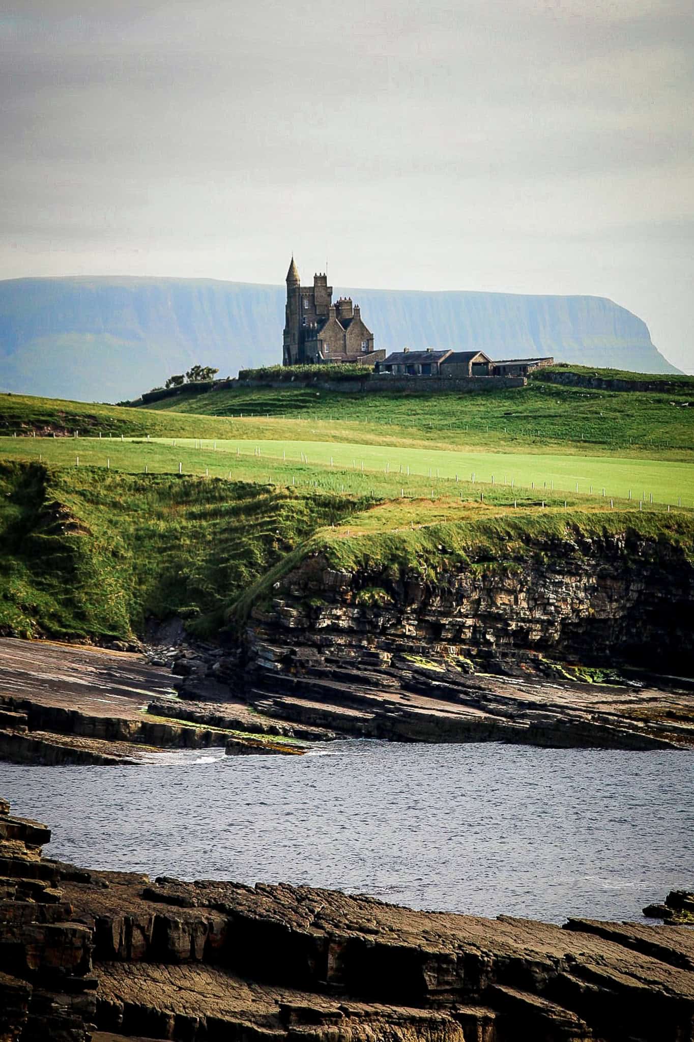 A castle-like structure on a grassy hill, surrounded by rolling cliffs and a rugged coastline. The foreground features layered rock formations beside a calm body of water.