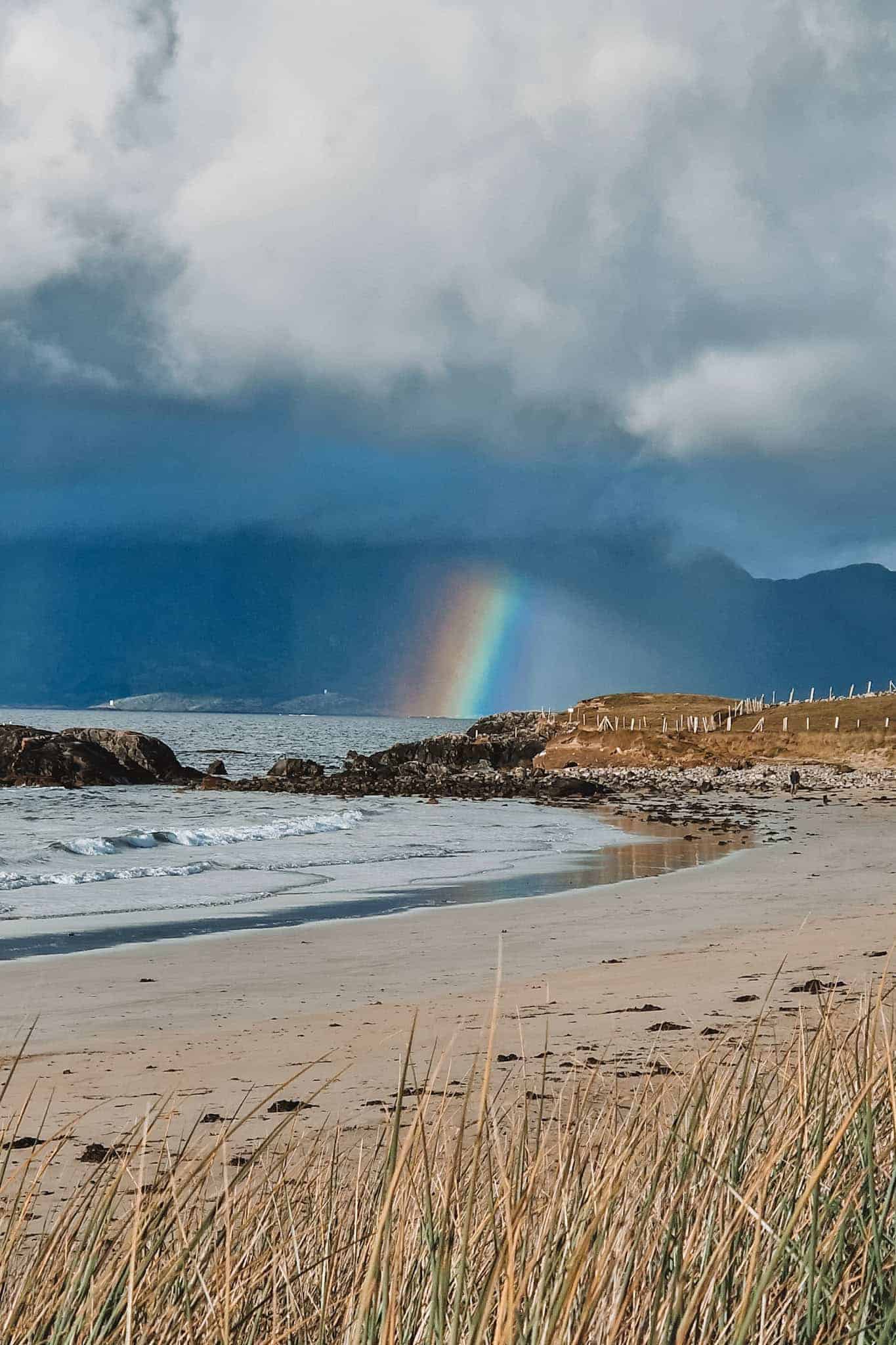 A vibrant rainbow arches across the sky over the coastline of Connemara, with a sandy beach and grassy dunes in the foreground.