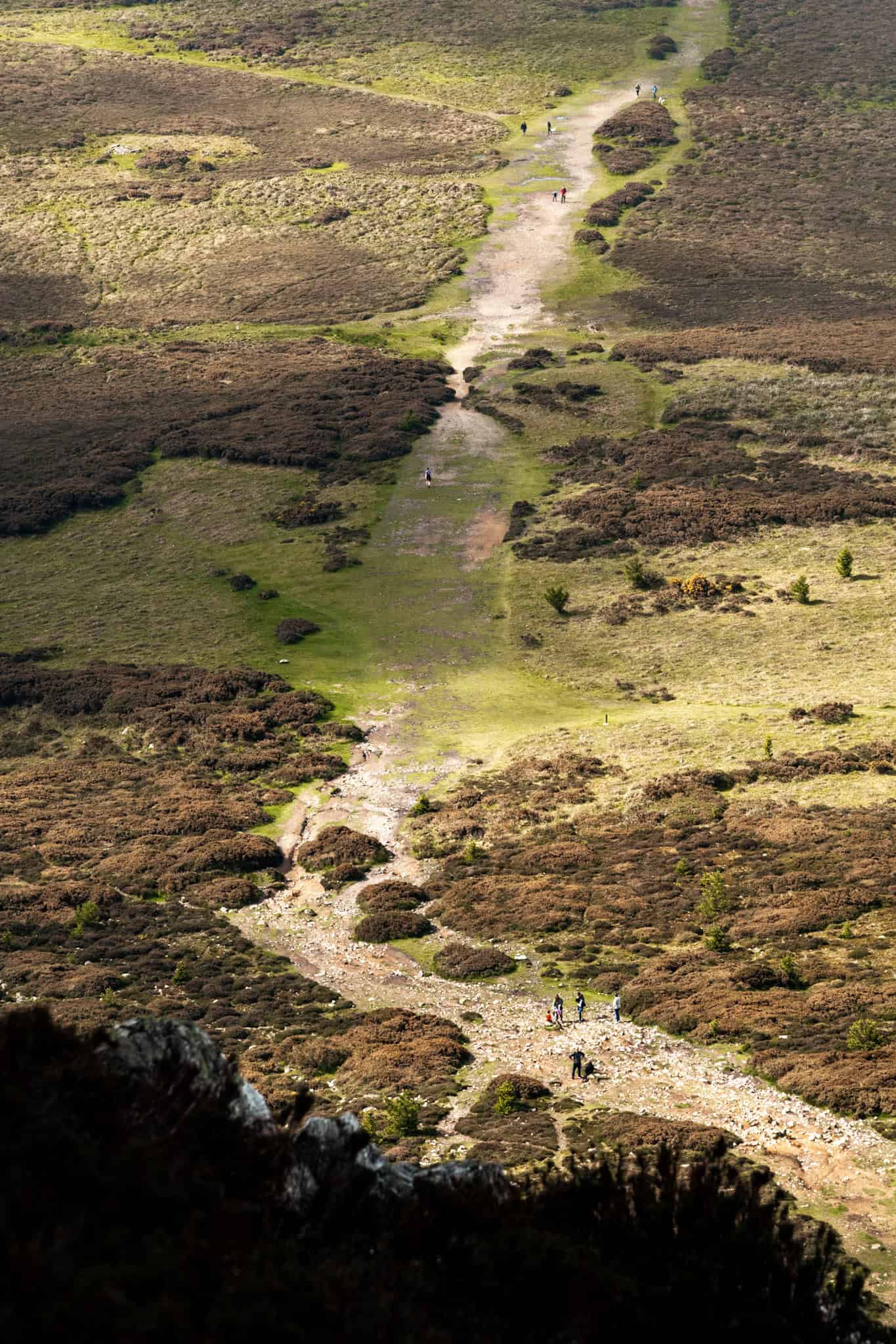 A narrow path winding through a rugged landscape of green and brown fields, with hikers walking along the trail surrounded by shrubs and grass.