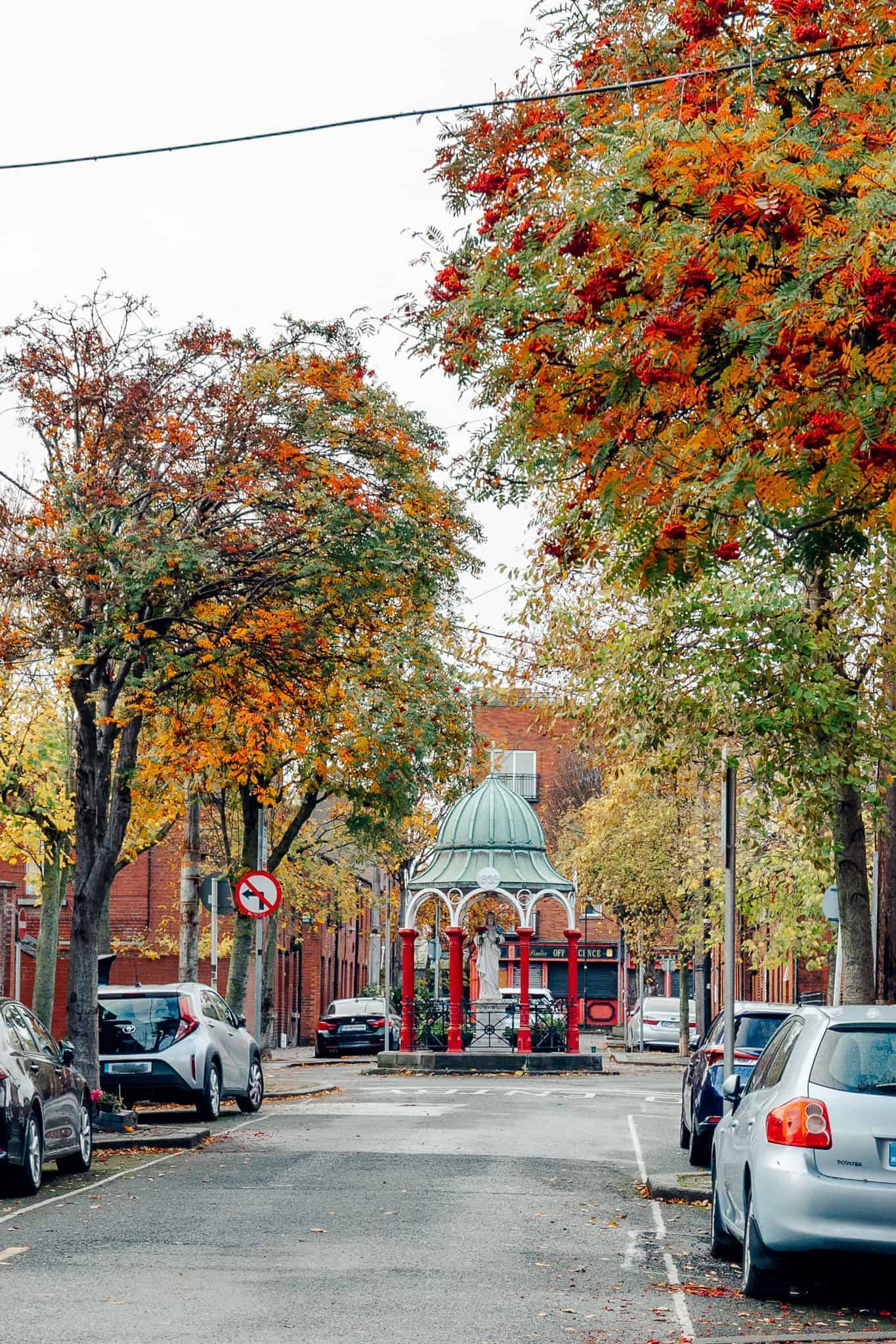 A quiet city street framed by trees with autumn foliage and parked cars. At the center of the image, a charming gazebo with a green dome and red columns stands as a focal point.