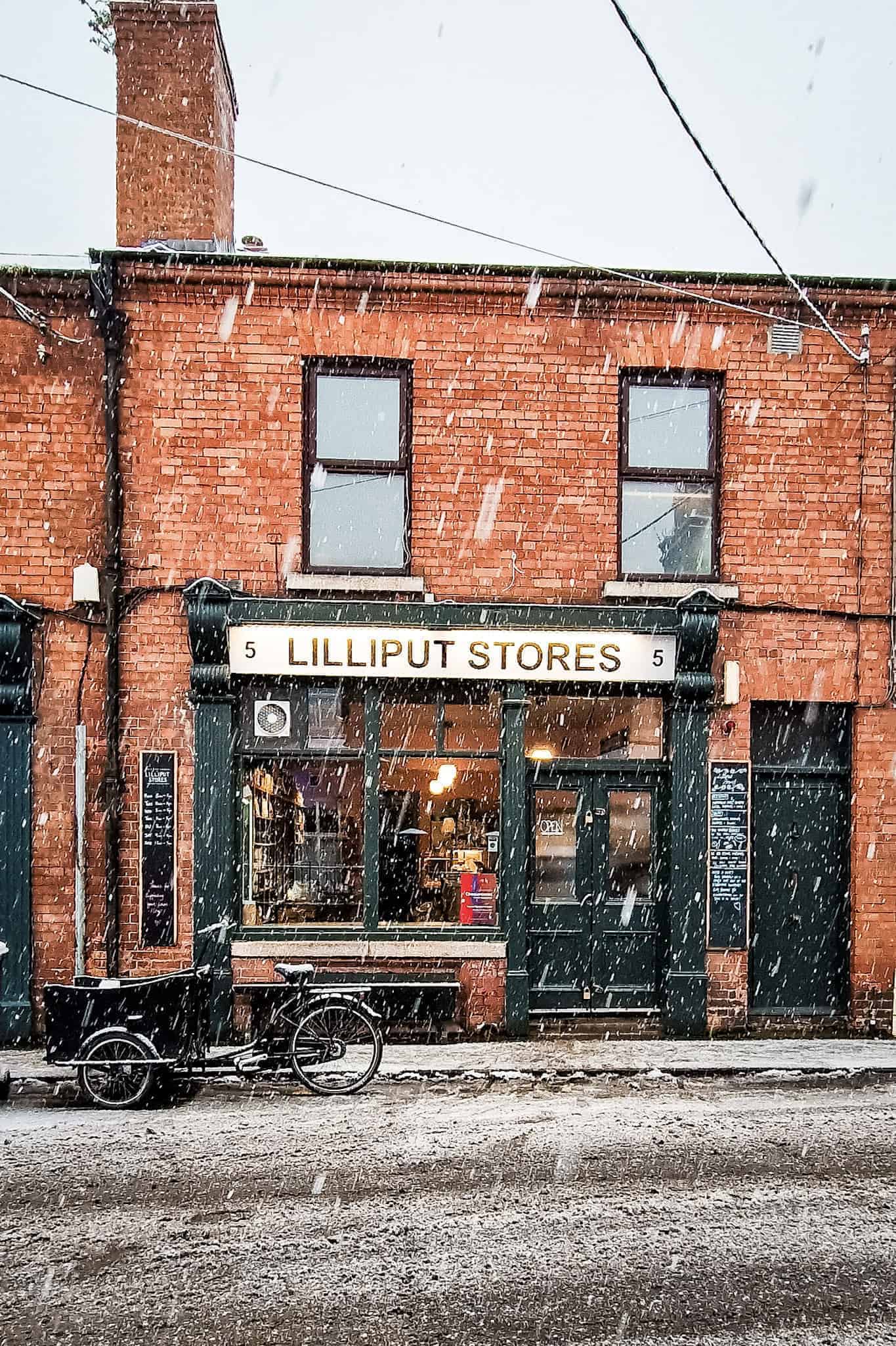 A charming red-brick storefront labeled "Lilliput Stores," framed by heavy snowflakes falling against the wintry backdrop. A bicycle is parked in front, adding to the quaint, picturesque scene.