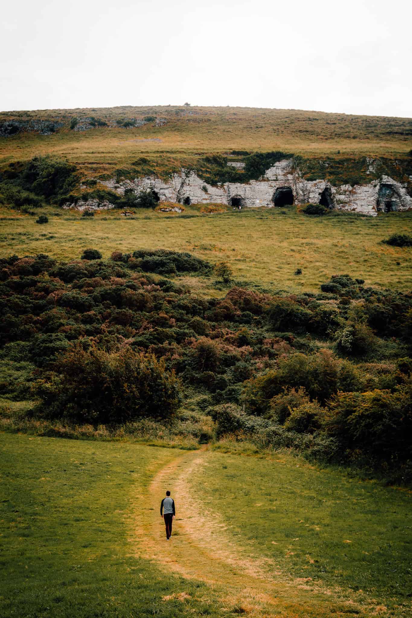 A person walking along a dirt path through an open, grassy field, with rocky caves embedded in a hillside ahead. The rugged terrain is surrounded by lush vegetation.