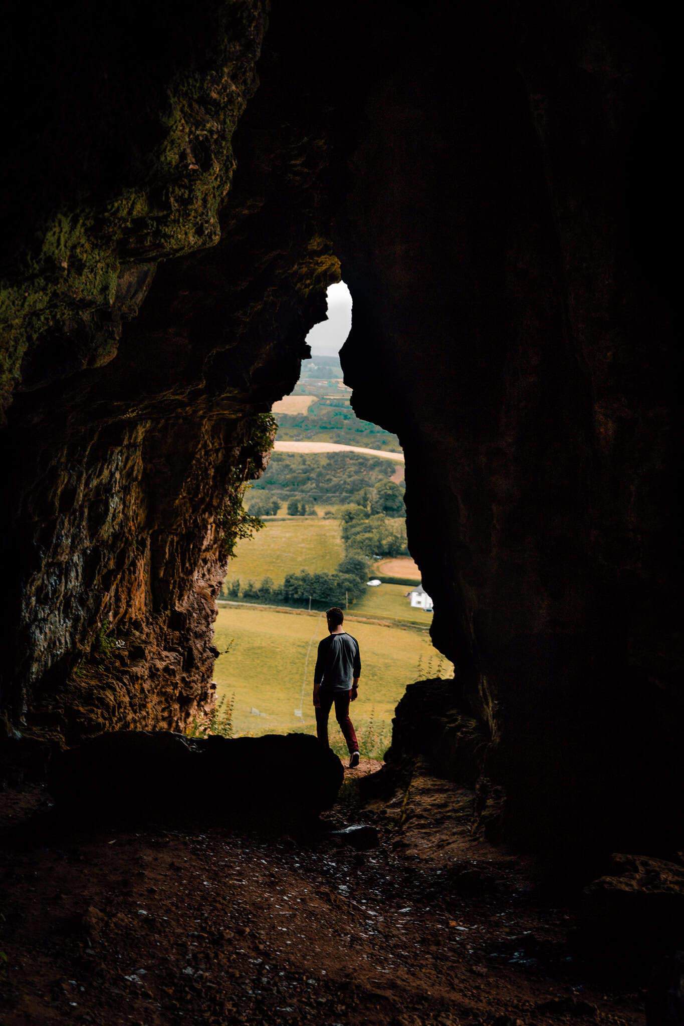 A person silhouetted against the entrance of a dark cave, looking out over a bright, green countryside with fields and trees. The jagged cave walls frame the pastoral view.