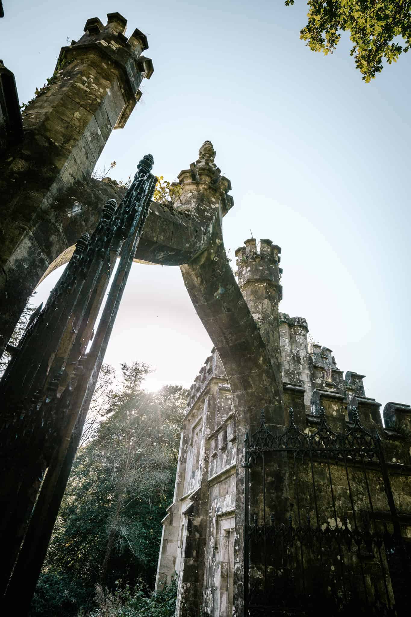 A low-angle view of the ornate, arched stone gate of Ballysaggartmore Lodge, framed by two towering structures with battlements. Sunlight peeks through the trees in the background, casting a soft glow on the weathered stone and iron gate.