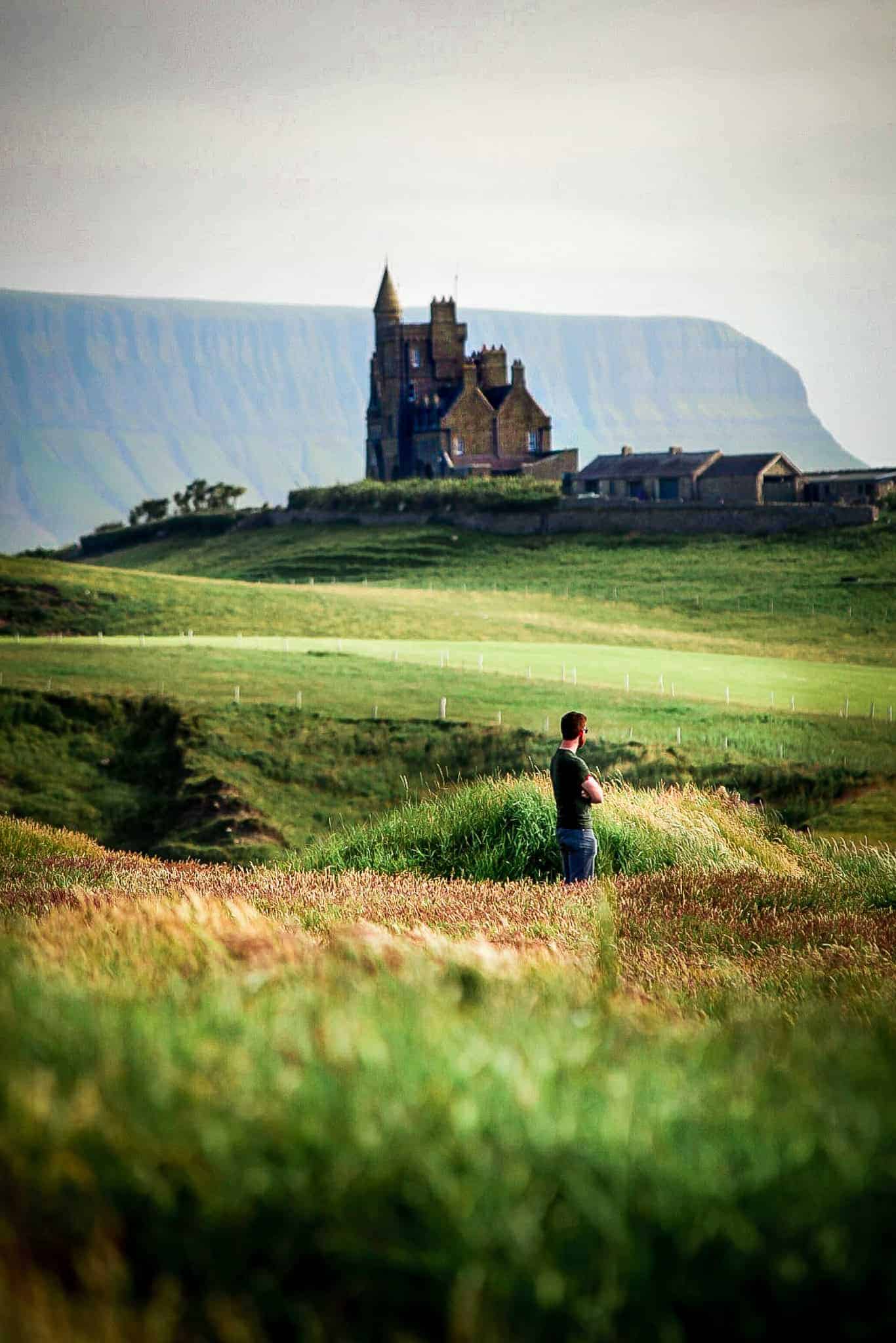 A lone man stands in a windswept field, gazing toward a dramatic, castle-like structure perched on a grassy hilltop. In the background, sheer cliffs rise against a cloudy sky.