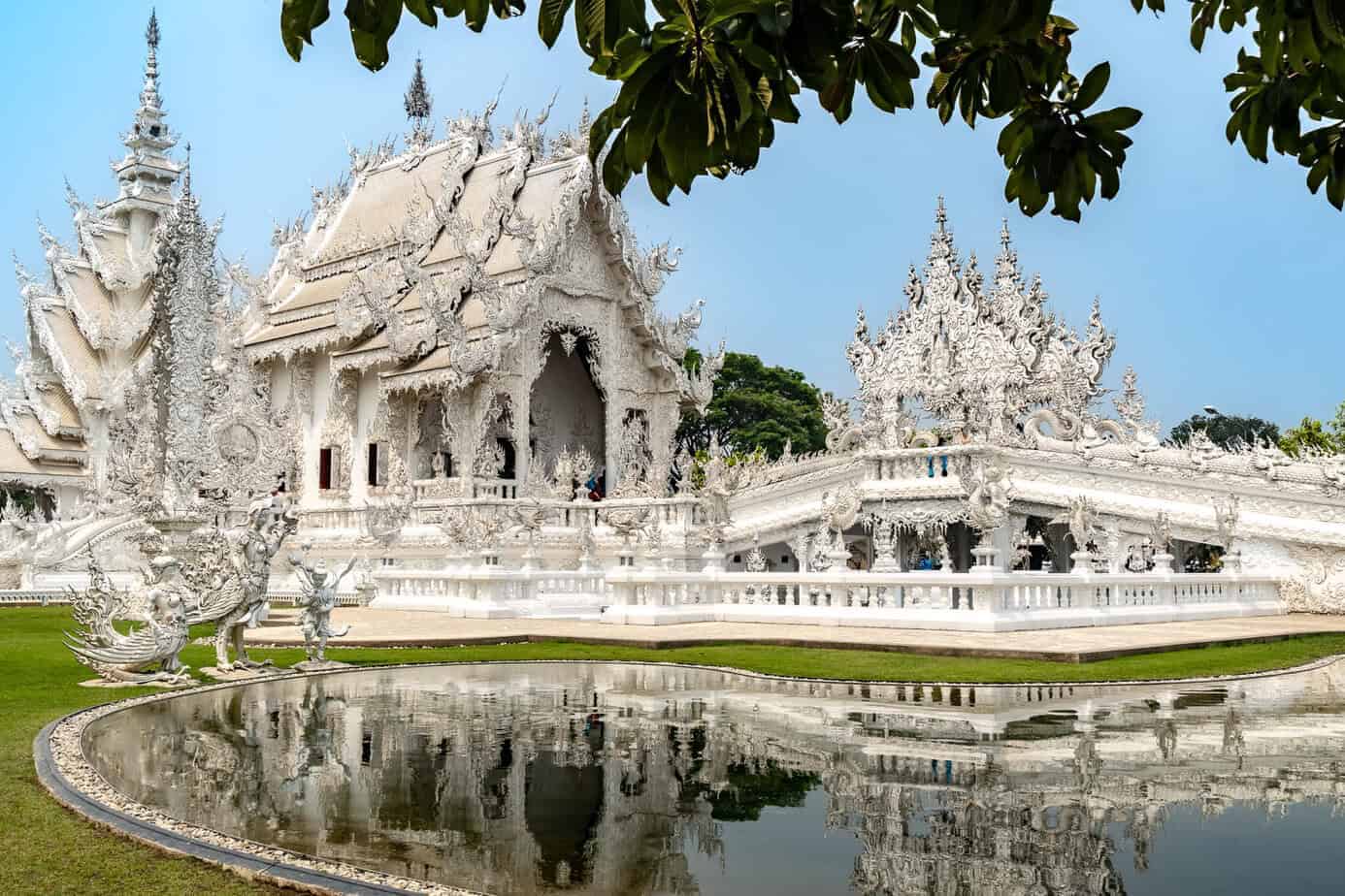 Chiang Rai: White Temple, Geheimtipps & Highlights im Norden Thailands 4 Der kunstvoll verzierte White Temple mit Spiegelmosaiken, der sich im stillen Teich spiegelt, umgeben von Grün und überhängenden Ästen unter einem klaren blauen Himmel.