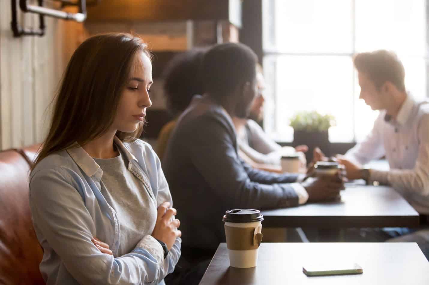 A sad woman with crossed arms in a coffee shop near other happy people thinking "I'll never find true love".