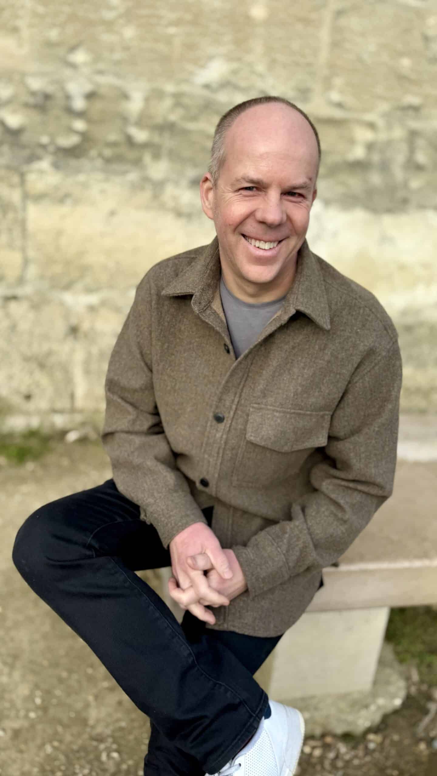 Derek Fredrickson, founder and owner of The COO Solution, sitting outside on a bench with his legs crossed an dhis hands clasped in his lap, smiling and looking at the camera