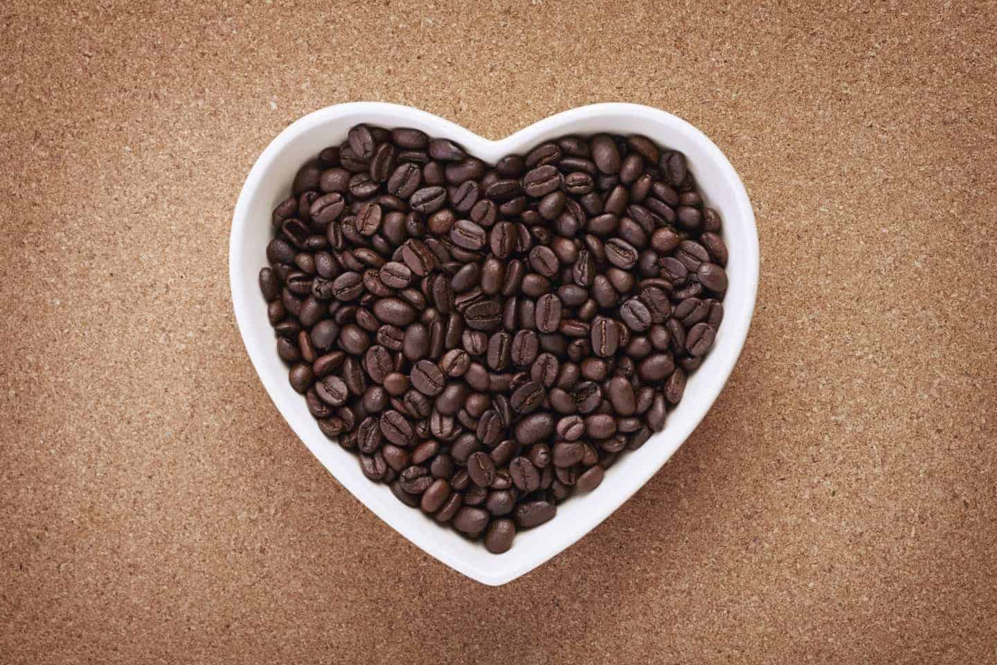 A white heart shaped bowl on wooden surface, filled with coffee beans