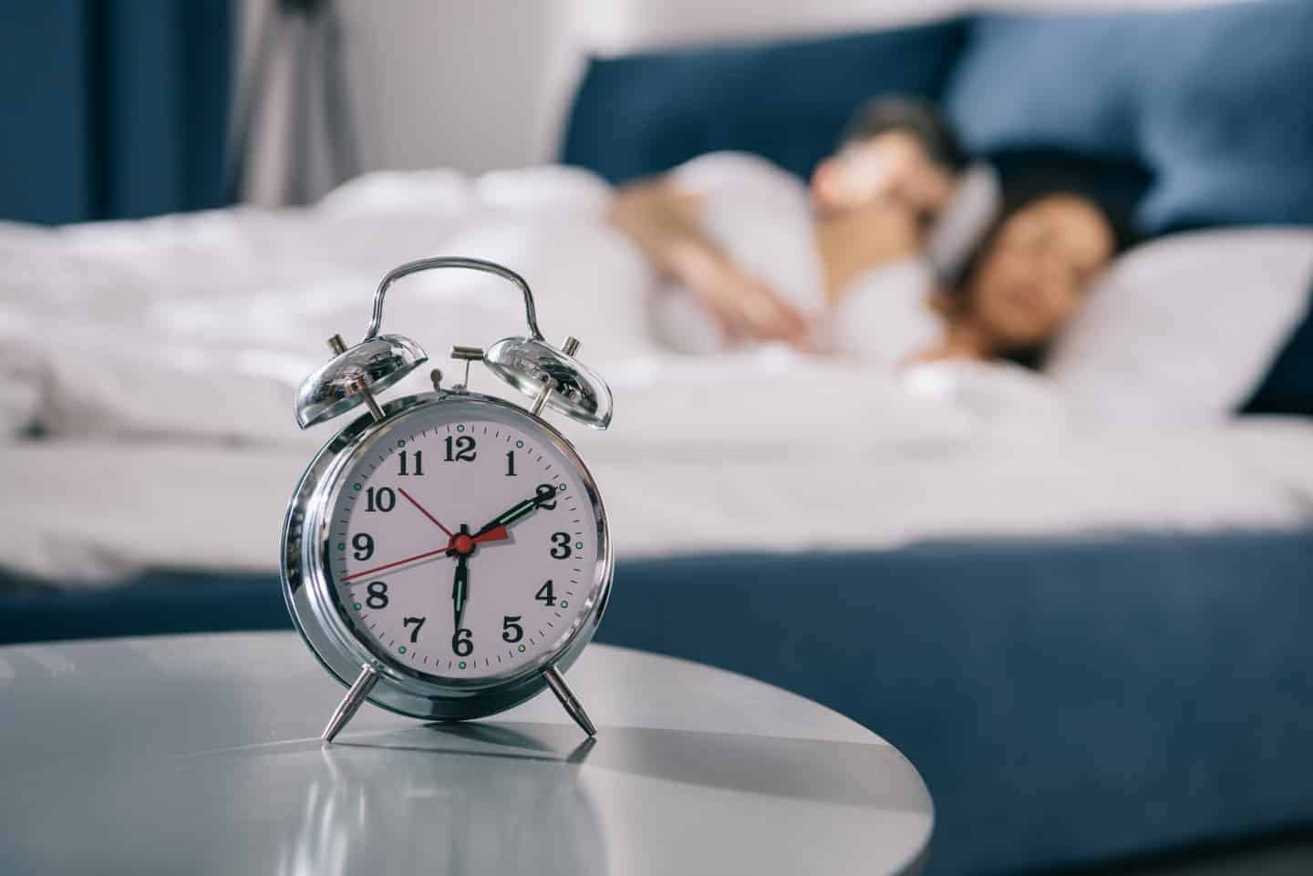 A clock that is sitting on a table with a couple in bed in the background