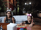 Duas meninas com tiaras luminosas, uma de orelhas de coelho e outra de laço rosa, sentadas em uma mesa de madeira à noite, com a mais nova sor