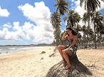 Uma mulher sentada em um tronco de palmeira na praia, protegendo os olhos do sol e olhando para o mar, com muitas palmeiras e céu azul ao fundo.