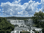 Vista panorâmica das Cataratas do Iguaçu, exibindo múltiplas quedas d'água imponentes em meio à floresta exuberante e um céu azul com nuvens.