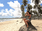 Uma mulher sentada em um tronco de palmeira na praia, protegendo os olhos do sol e olhando para o mar, com muitas palmeiras e céu azul ao fundo.