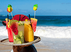 Uma pessoa carrega uma bandeja com quatro bebidas tropicais coloridas, decoradas com frutas, em uma praia ensolarada com o oceano azul ao fundo.