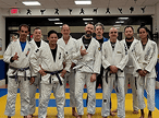 Nove praticantes de jiu-jítsu, a maioria vestindo quimonos brancos com faixas roxas e pretas, posam em um dojo com tatames azuis e amarelos.