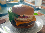 Um hambúrguer gourmet alto e empilhado com carne, queijo derretido, ovo frito, tomate, cebola e alface, servido em um prato de vidro.
