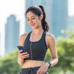 Uma jovem mulher asiática sorridente, vestindo top esportivo cinza e fones de ouvido, olha para o celular ao ar livre com prédios urbanos desfocados no fundo.