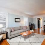Sun-drenched living room at 3764 Alabama in North Park with a grey sofa, hardwood floors, a geometric rug, and large windows with white blinds.