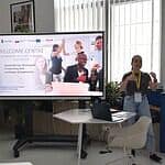Uczestniczka prezentuje podczas PHD RISK Summerschool, obok ekran z informacją o warsztatach, w nowoczesnym biurze z dużymi oknami i naturalnym oświetleniem.