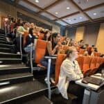 Grupa studentów i naukowców uczestniczących w PHD RISK Summerschool podczas wykładu w nowoczesnej sali konferencyjnej.