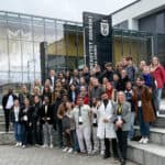 Grupa uczestników PHD RISK Summer School przed budynkiem Uniwersytetu Gdańskiego, z logo uczelni i nowoczesną architekturą w tle, podczas wydarzenia naukowego.