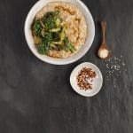 A bowl of savoury porridge topped with sautéed greens and sauce. A small dish of red pepper flakes and a spoon with sesame seeds are on the side, set on a dark slate surface. Perfect for those seeking unique savoury porridge ideas.
