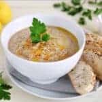 A white bowl of aubergine soup with bread on the side of the plate