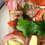 Close-up of grilled meat and vegetable skewers on a plate, garnished with parsley, with a sauce dish in the background.