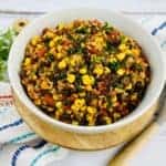 A bowl of vibrant tex mex salad with mixed corn, black beans, tomatoes, and herbs sits on a wooden board. A patterned napkin and spoon lie beside it, inviting a delicious dive into this quinoa-infused delight.