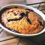 A freshly baked berry cobbler, reminiscent of Slimming World baked oats, sits in an oval dish on a metal cooling rack. It has a golden-brown crust with purple berry filling peeking through cracks, all set against a wooden countertop.
