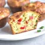 Savory breakfast egg muffins with visible ingredients like chopped vegetables and meat are arranged on a white plate. One muffin is sliced in half, revealing its texture and delicious filling inside.