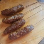 Four grilled skewers of meat sit enticingly on a round wooden cutting board, reminiscent of a five-star butcher review.