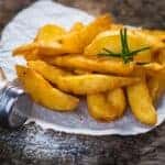 potato wedges on a white plate with a sprig of rosemary and spilt salt on wooden table
