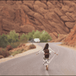 Woman with backpack walking down a winding desert road, arms spread wide, red rock hills in the background