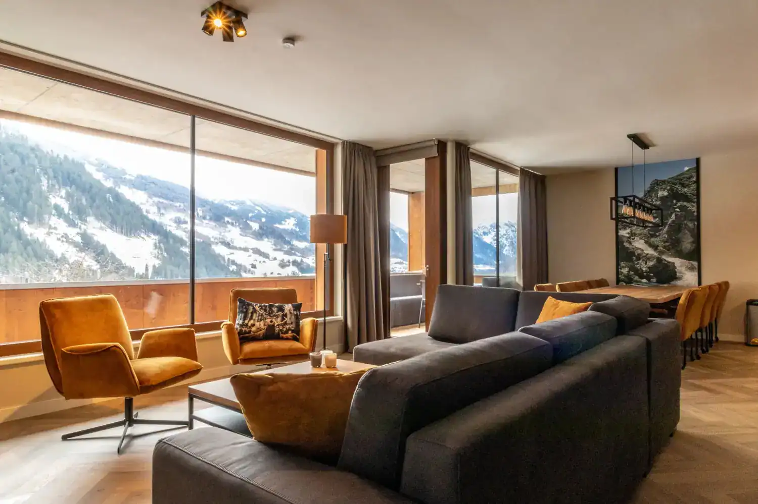 Gemütlicher Wohnbereich der Silvretta Panorama Suites mit dunkelgrauem Sofa, senfgelben Sesseln und deckenhohen Panoramafenstern, die eine weite Aussicht auf die verschneiten Berge bieten.