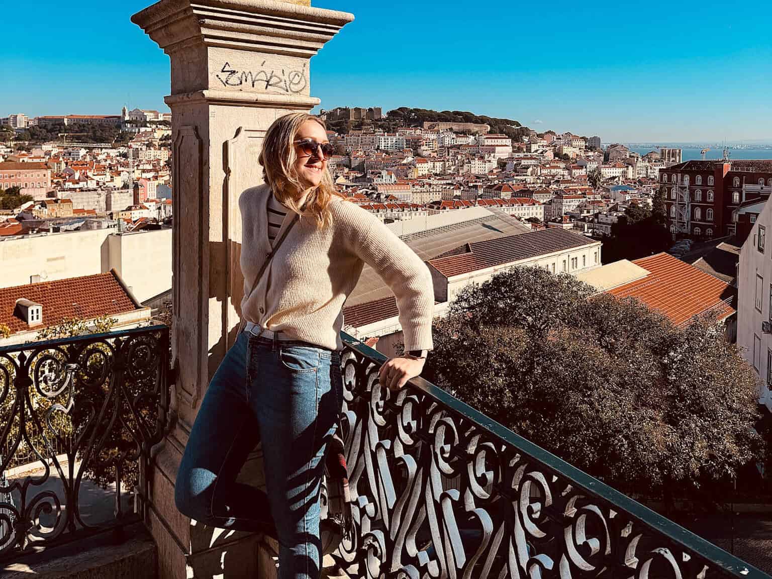 Woman in a cream cardigan and blue jeans standing on a balcony overlooking Lisbon, Portugal, with a decorative stone column and wrought-iron railing in the foreground. Travelling with ADHD doesn't have to limit your experiences and enjoyment if you plan, manage and adapt.