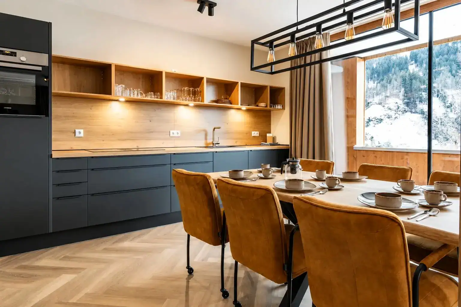 Moderne, offene Küche in den Silvretta Panorama Suites von UplandParcs mit dunkelgrauen Schränken, großem Holzesstisch, senfgelben Stühlen und Blick durch das Fenster auf eine verschneite Berglandschaft.