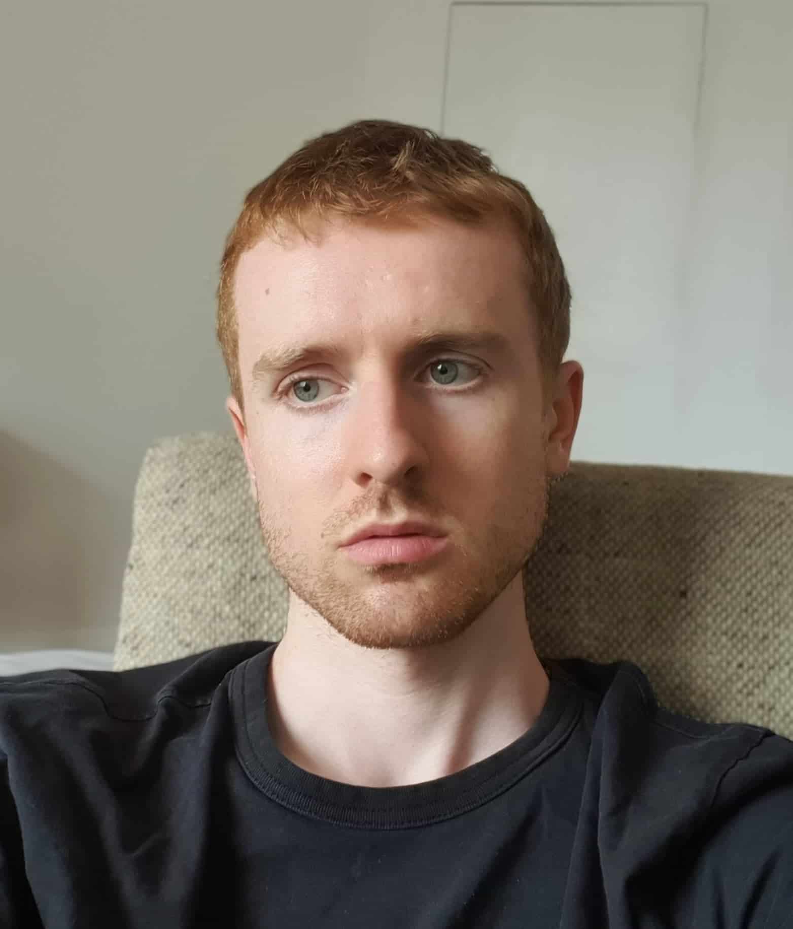 Young man with red hair and blue eyes relaxing on a sofa, casual black T-shirt, natural indoor lighting, modern interior, lifestyle, portrait, comfortable home setting.