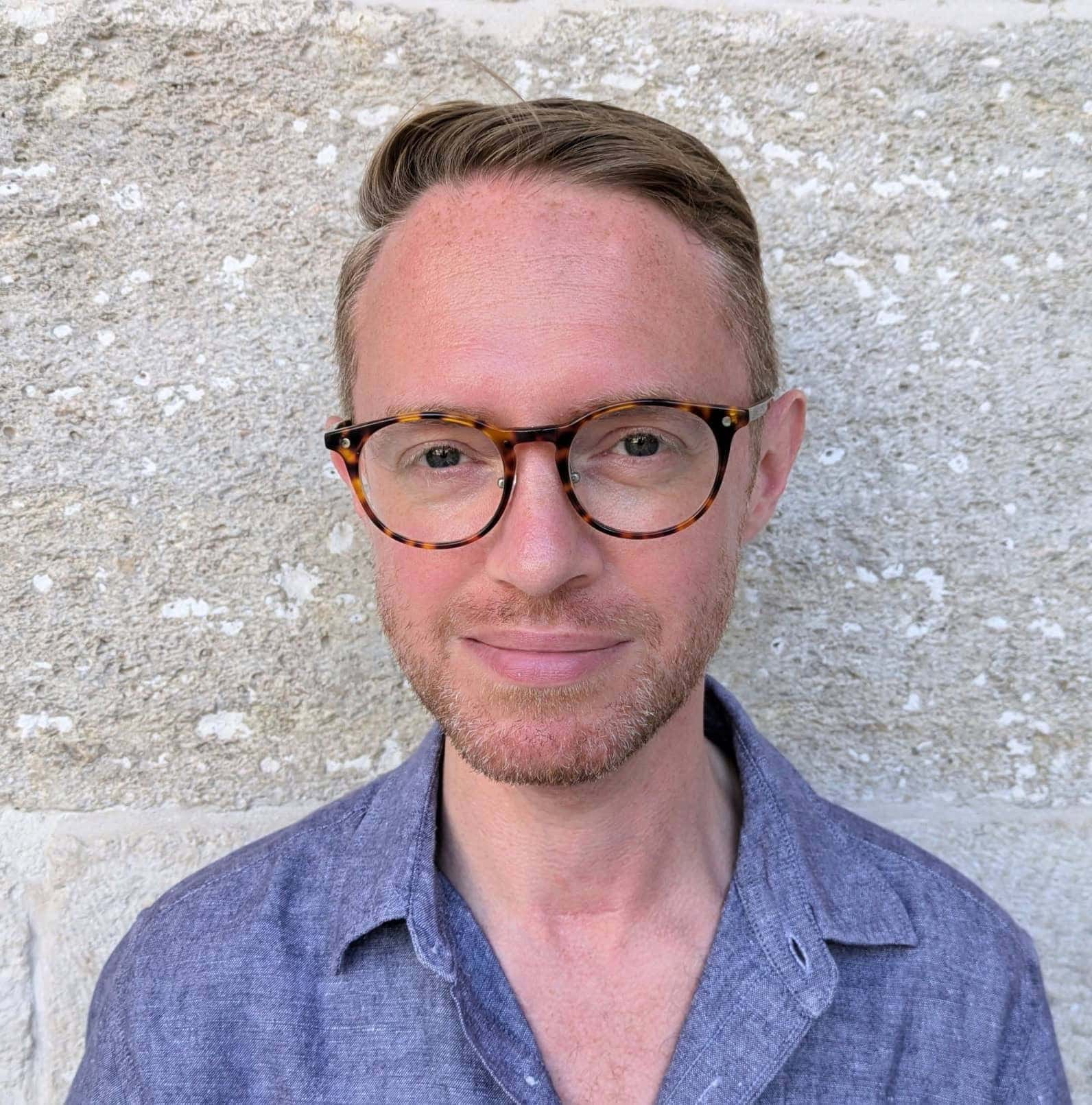 Portrait of a young man wearing tortoiseshell glasses and a blue shirt, standing outdoors against a textured stone wall, showcasing modern eyewear and fashionable casual style.