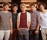 Cinco jovens da banda One Direction sorriem e posam casualmente com roupas diversas, em frente a um fundo escuro com fileiras de luzes brilhantes.