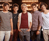 Cinco jovens da banda One Direction sorriem e posam casualmente com roupas diversas, em frente a um fundo escuro com fileiras de luzes brilhantes.
