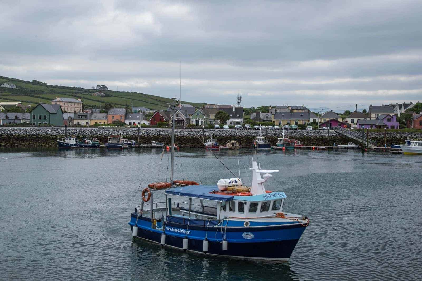 A small blue fishing boat named "Star of the Sea" floats in a calm harbor, with colorful houses and green hills in the background under a cloudy sky.