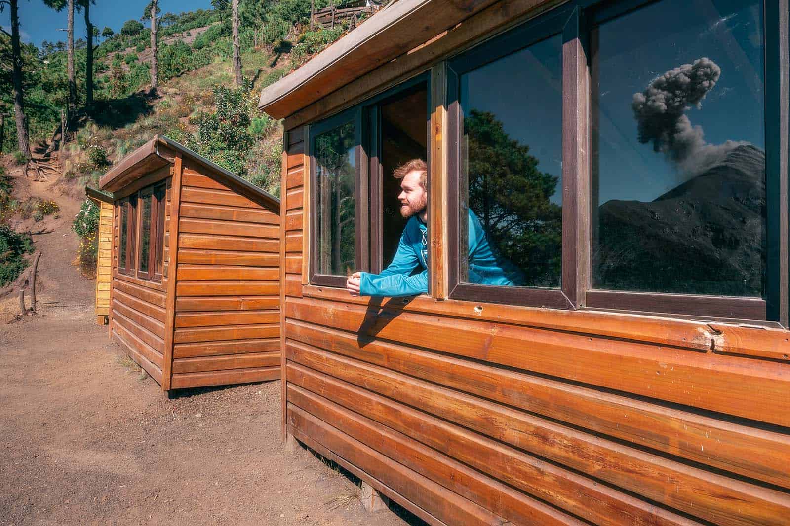 Guest leaning out of an OX Expeditions cabin window at Acatenango campsite watching Fuego volcano erupt