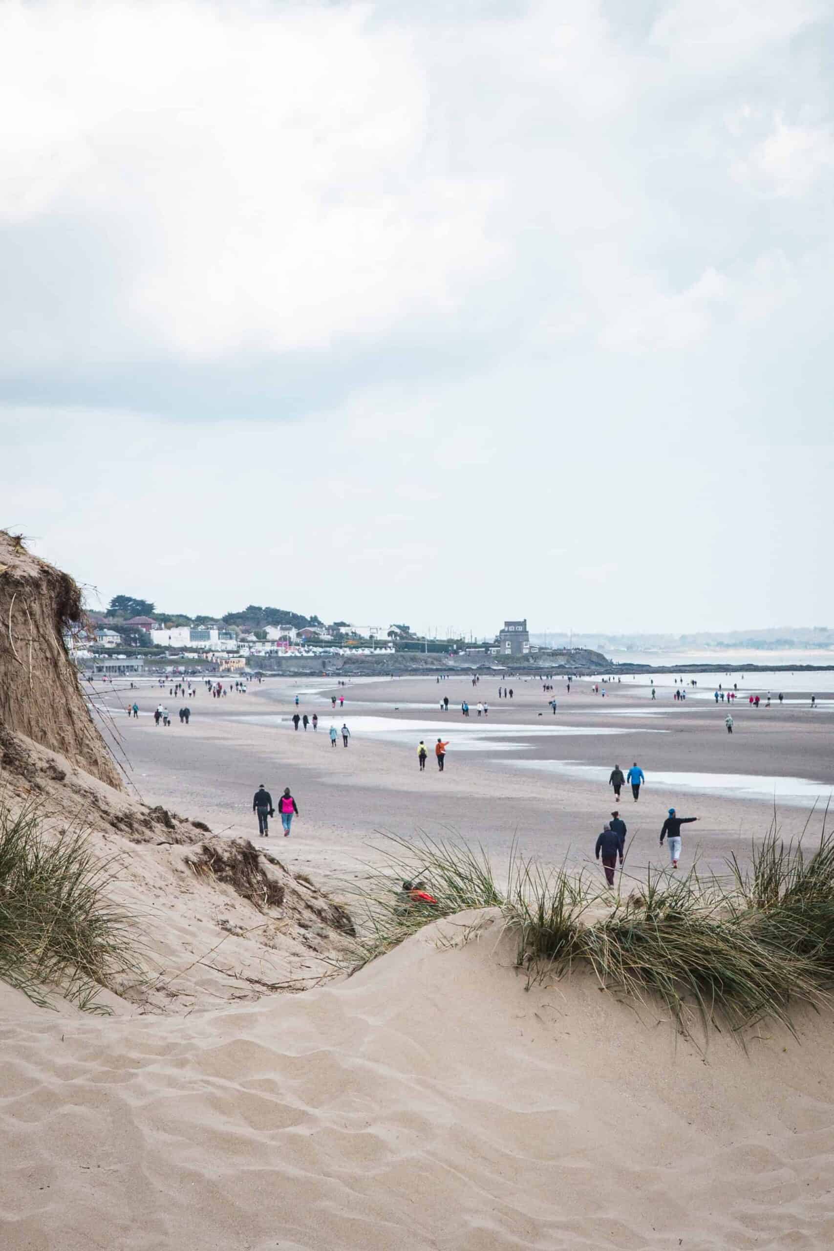 A sandy beach at Portmanock, Ireland, with people walking along the shore. The scene includes distant houses and a cloudy sky.