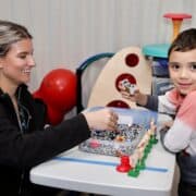 A woman and a young boy sit at a table playing with toys and sensory beads. The woman smiles at the boy, who looks toward the camera while holding a toy car. Colorful play equipment is visible in the background.