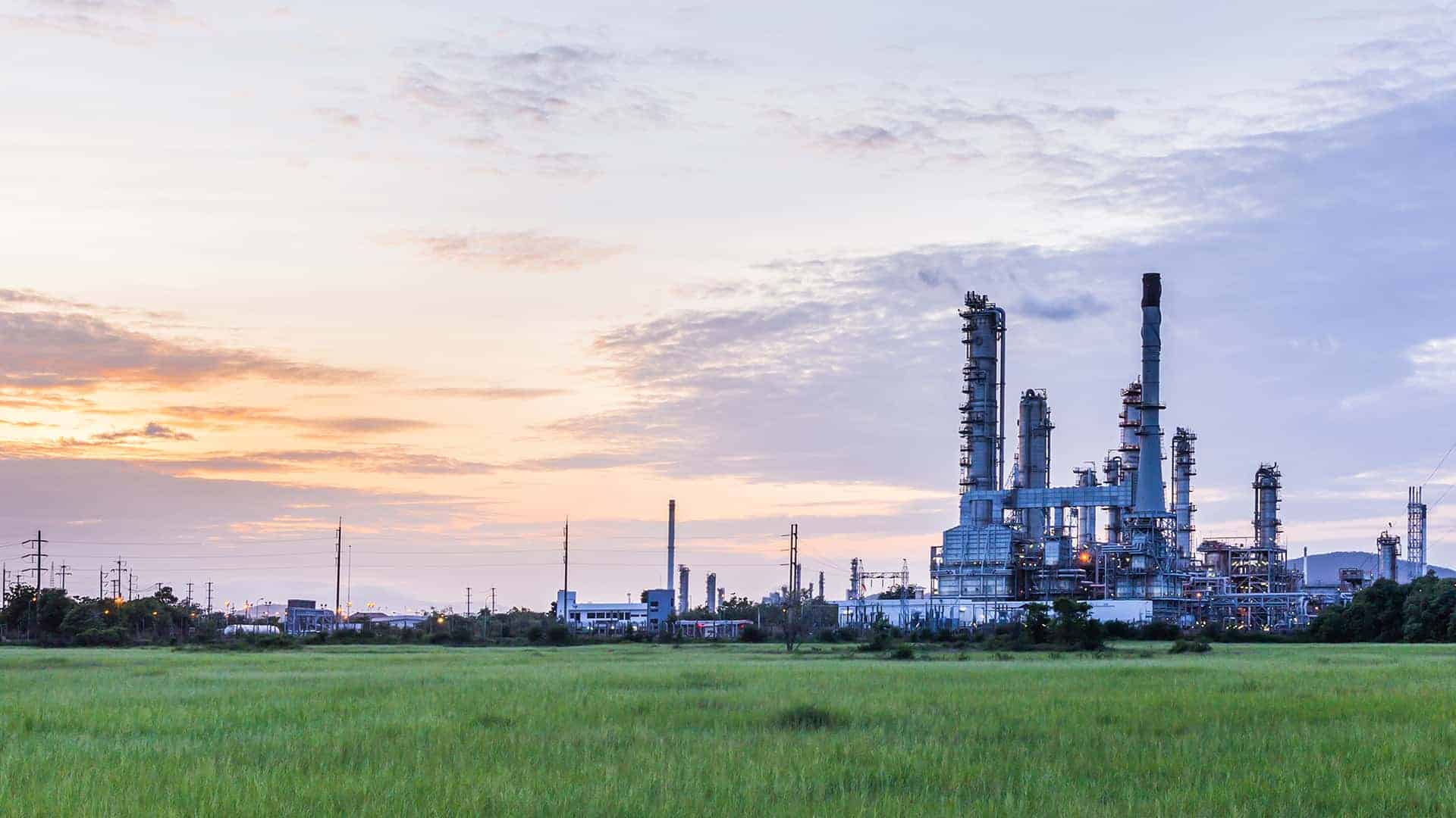 Oil refinery complex at dusk with green fields in the foreground, industrial towers and processing equipment silhouetted against a pastel sunset sky — representing the energy sector facilities affected by the Clean Power Plan debate.